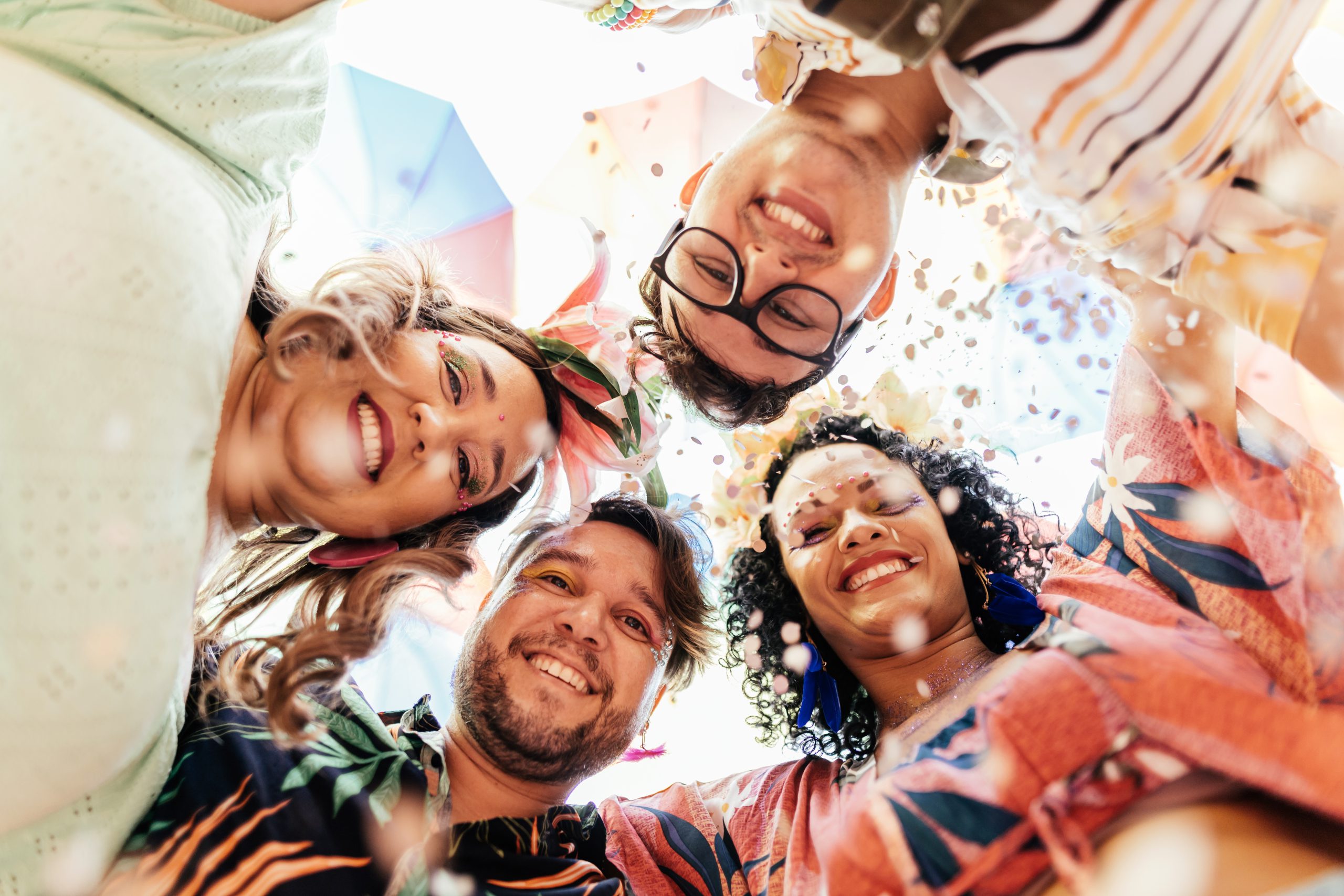 Grupo de pessoas celebrando Carnaval ao ar livre, vistos de baixo para cima, rodeados por confetes coloridos e luz natural, transmitindo alegria e energia festiva.